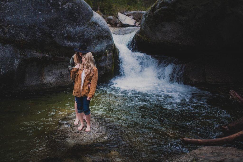 Engagement photography in Sequoia National Park of a stylish, adventurous couple posing on a rock in the middle of a creek wtih a waterfall in the background.