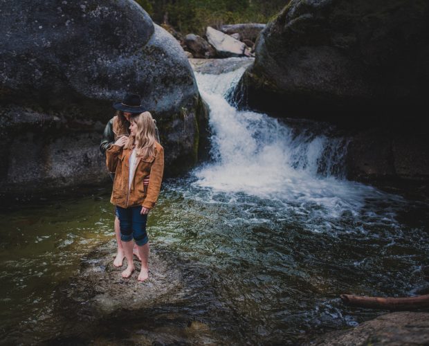 Engagement photography in Sequoia National Park of a stylish, adventurous couple posing on a rock in the middle of a creek wtih a waterfall in the background.