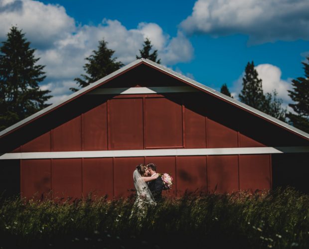 Outdoor wedding photography of a bride and groom posing in front of a red barn on a farm.