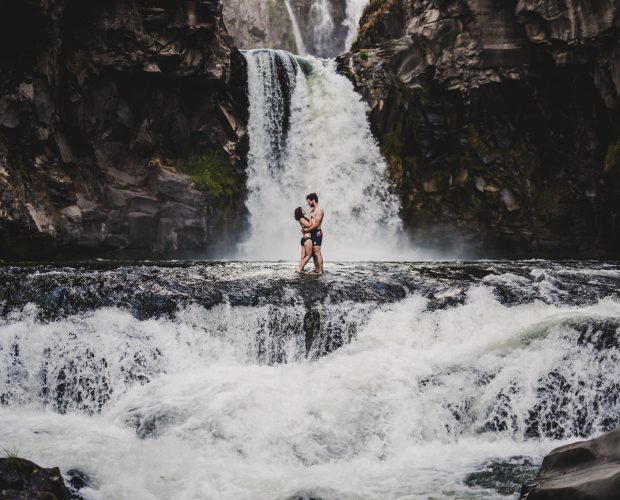 An engaged couple stands on the edge of a waterfall at White River Falls in Tygh Valley, Oregon, during their engagement session photography by Portland, Oregon, destination wedding photographer Kyle Carnes.