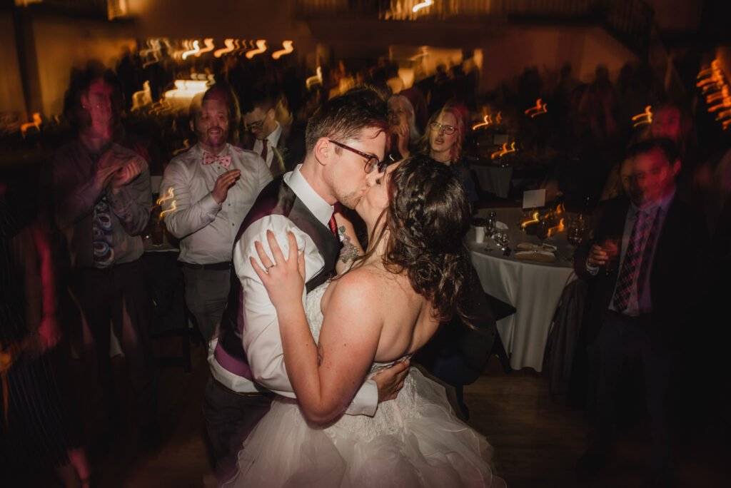 A couple kisses on the dance floor at a lively wedding reception, surrounded by blurred guests, capturing a moment for their wedding photos at The Evergreen.
