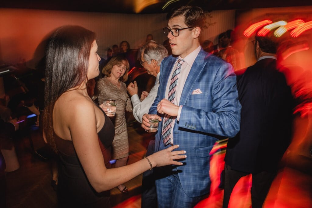 A man in a blue suit and a woman in a black dress are talking at a lively party with blurred lights in the background, perhaps discussing the wedding photos at The Evergreen.