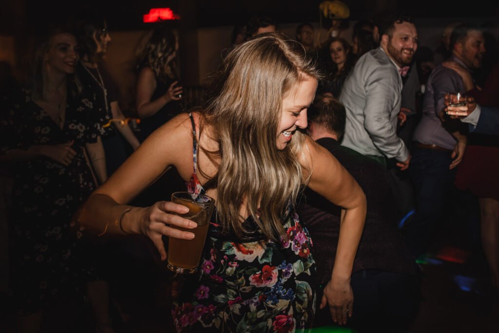 At a lively party, a woman in a floral dress joyfully dances with a drink in hand. Guests around her are also enjoying and dancing, capturing moments fit for wedding photos at The Evergreen.