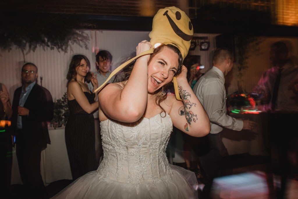 The bride, in a stunning white dress, laughs with a smiley face hat perched on her head at a lively wedding reception. Joyful moments like these make for perfect wedding photos at The Evergreen.