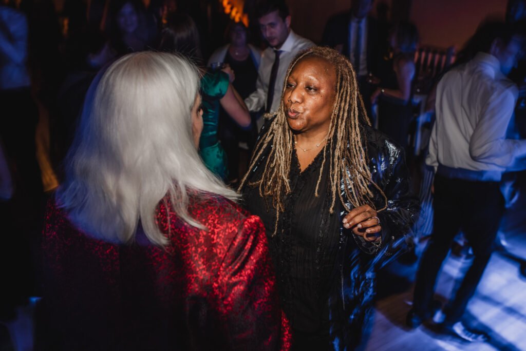 At a lively party, two people are deep in conversation, surrounded by guests swaying on a dimly lit dance floor. In the backdrop, memories are being captured with wedding photos at The Evergreen.