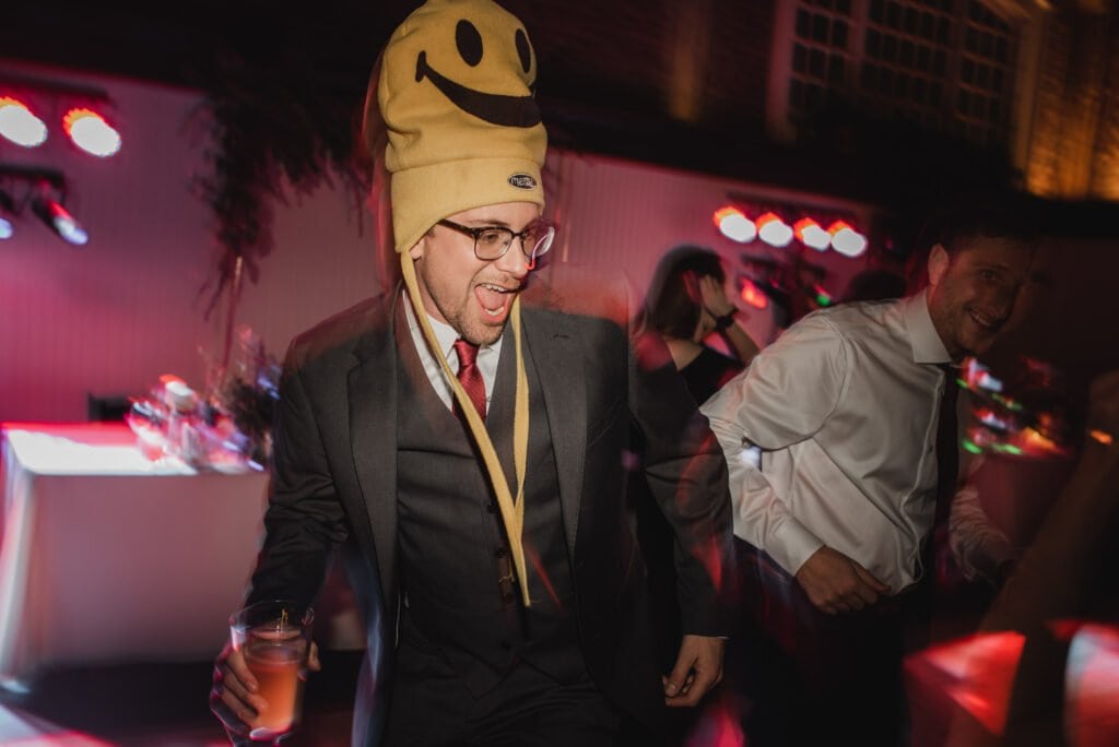 A person in a suit wearing a smiley face hat dances joyfully, drink in hand, with vibrant lights swaying around them. Its a scene that could easily fit among wedding photos at The Evergreen.