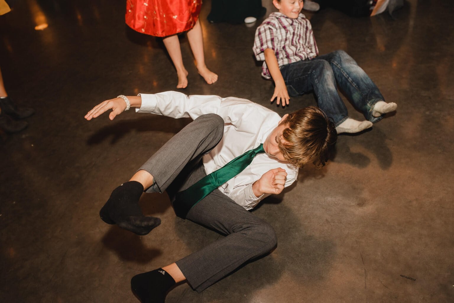 A boy in a white shirt and green tie playfully rolls on the floor at an indoor gathering, adding a candid moment to the wedding photos at Castaway Portland.