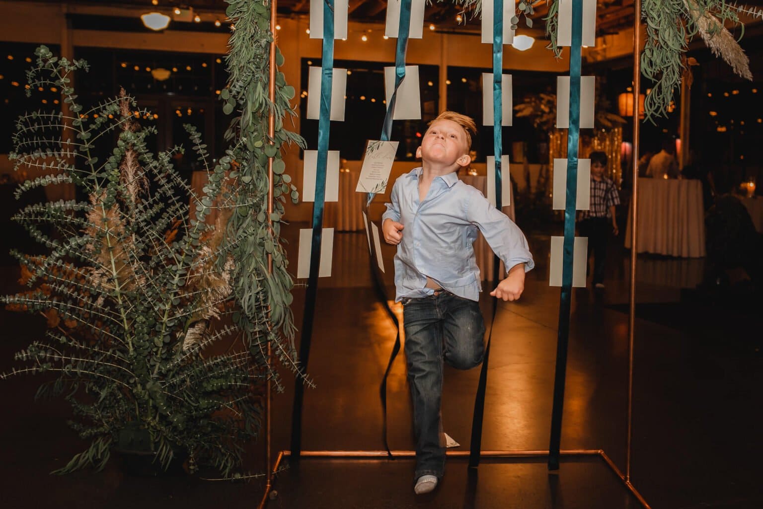 A boy playfully poses under a decorative arch adorned with greenery and hanging ribbons in a dimly lit venue, perfect for capturing wedding photos at Castaway Portland.