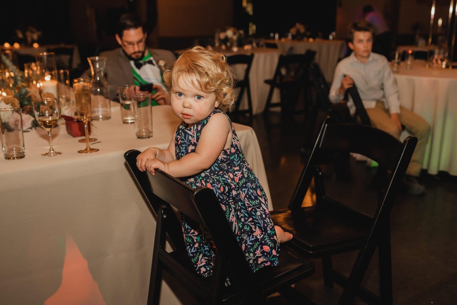 A toddler in a floral dress sits on a chair, looking at the camera. Two adults are in the background, seated at a beautifully decorated table, capturing a moment of sweetness during the wedding photos at Castaway Portland.
