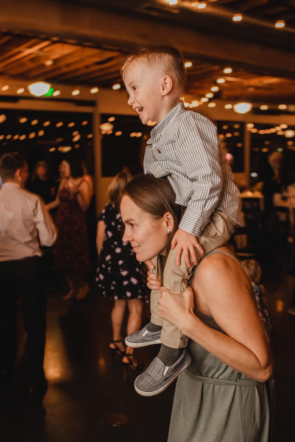 A woman carries a smiling child on her shoulders at a lively indoor event, capturing joyful moments of people dancing in the background during wedding photos at Castaway Portland.