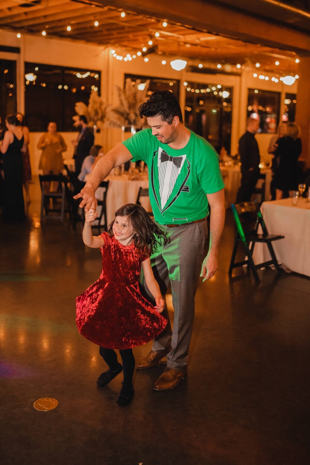 At a festive indoor event adorned with string lights, a man in a green tuxedo t-shirt dances joyfully with a girl in a red dress. The moment captures the lively spirit often seen in wedding photos at Castaway Portland.