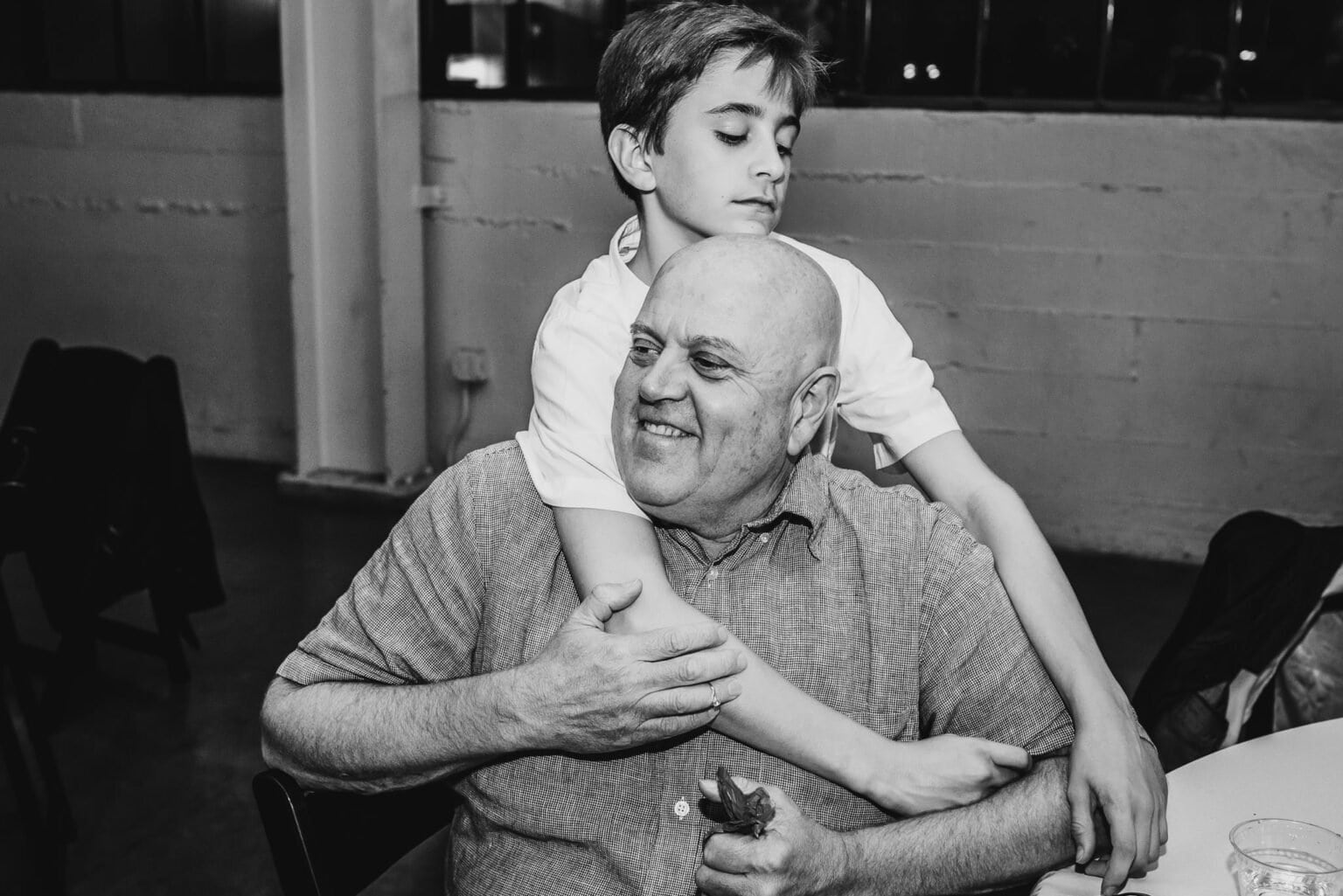 In a warmly lit room, a boy joyfully hugs an older man from behind, both sharing bright smiles.
