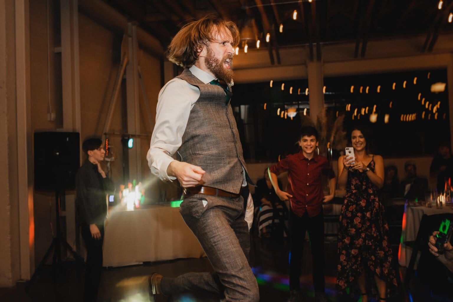 A man in a vest is joyfully dancing at an indoor event, captivating onlookers with vibrant lights in the background. Its like a scene straight out of wedding photos at Castaway Portland.