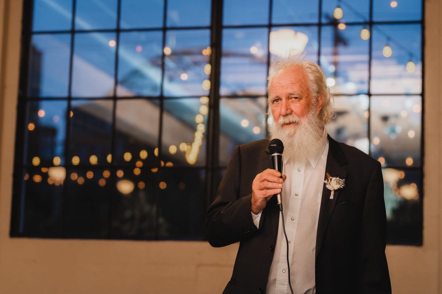 An elderly man with a white beard holds a microphone, speaking indoors with large windows and string lights behind him. This charming scene is perfect for wedding photos at Castaway Portland, creating timeless memories in an elegant setting.