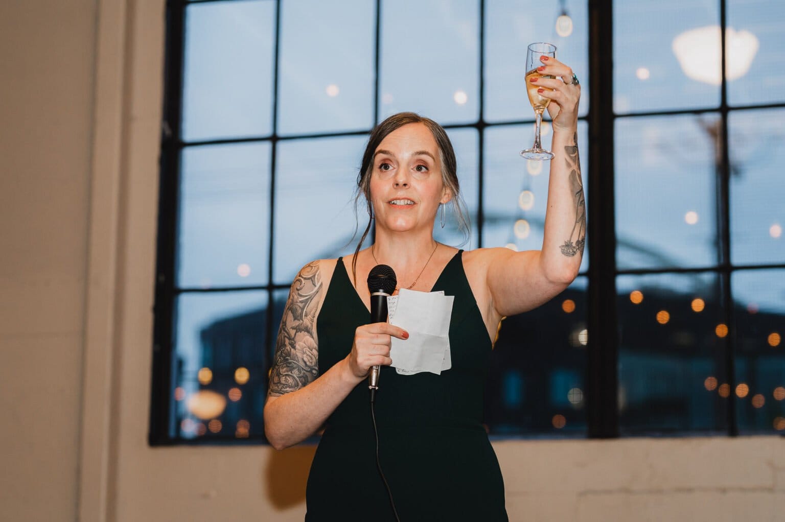 In a room with large windows at dusk, a person in a dark dress holds a microphone and raises a glass, capturing an unforgettable moment similar to those seen in stunning wedding photos at Castaway Portland.