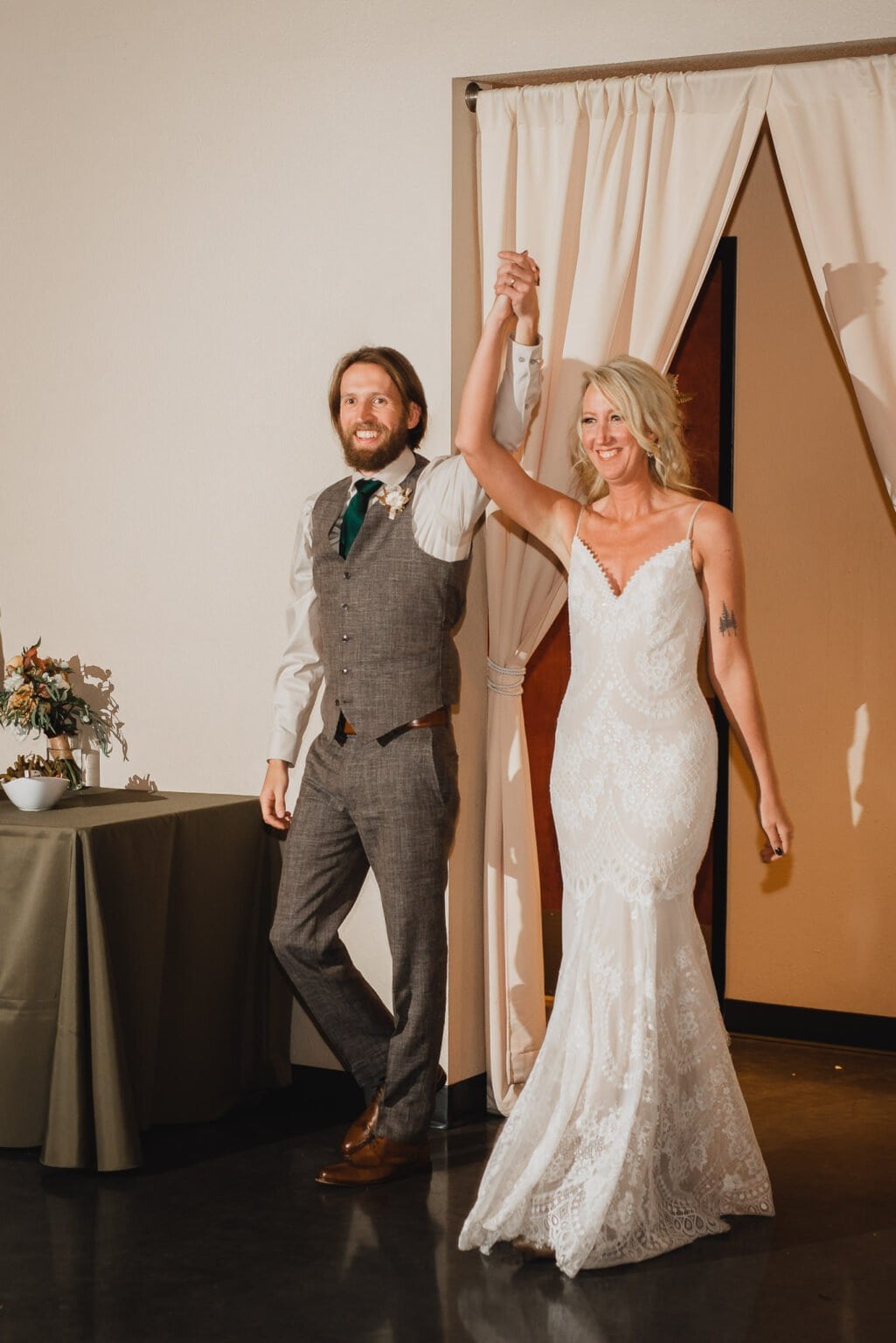 A smiling bride and groom hold hands, entering a room with a curtain backdrop and a small table with decorations, perfect for capturing wedding photos at Castaway Portland.