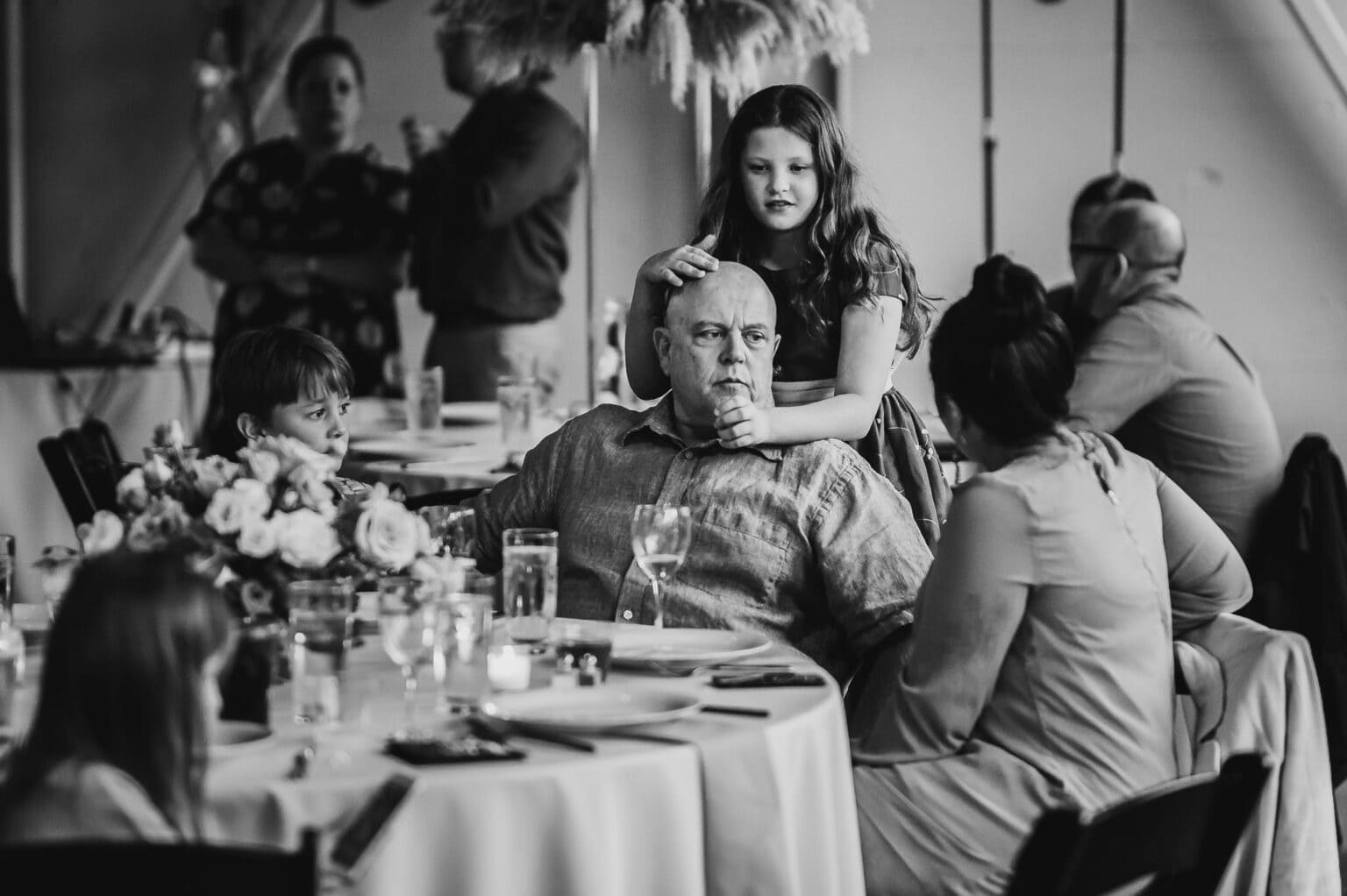 In a black and white photo, a girl rests her hands on an older mans head at a table in a bustling restaurant, surrounded by seated guests. Its one of the candid moments captured among the wedding photos at Castaway Portland.