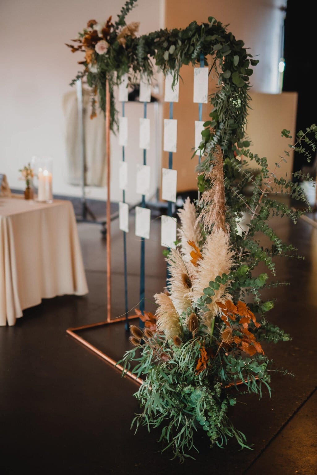 A wedding seating chart stand, embellished with greenery and pampas grass, is positioned near a draped table with candles, creating a picturesque scene perfect for wedding photos at Castaway Portland.