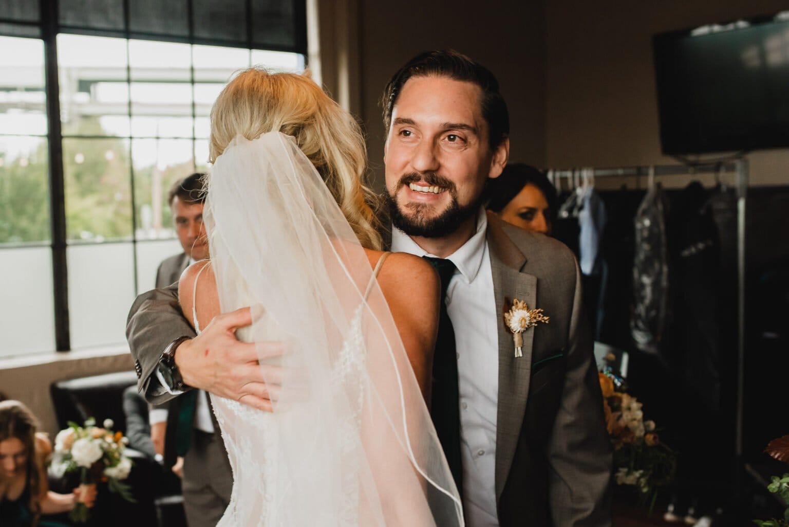 The groom in a suit embraces the bride in her white dress and veil indoors, surrounded by people and flowers. It captures the joy of wedding photos at Castaway Portland.