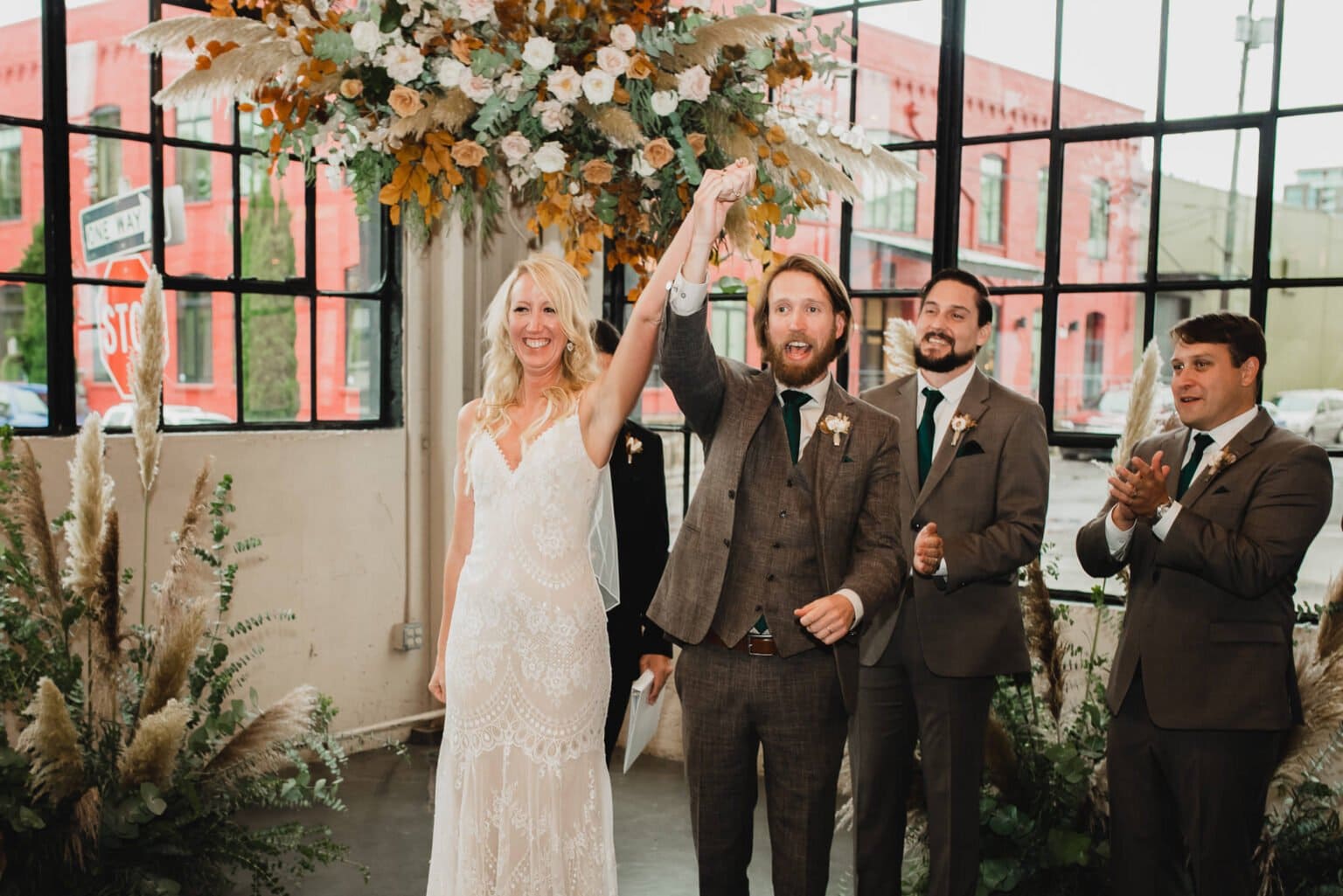 The bride and groom celebrate under a floral arch with three groomsmen clapping in a bright venue, highlighted by large windows, capturing stunning wedding photos at Castaway Portland.