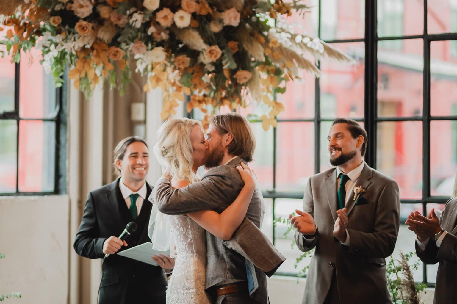 During their wedding ceremony at Castaway Portland, the bride and groom share a kiss as their guests clap and smile warmly.