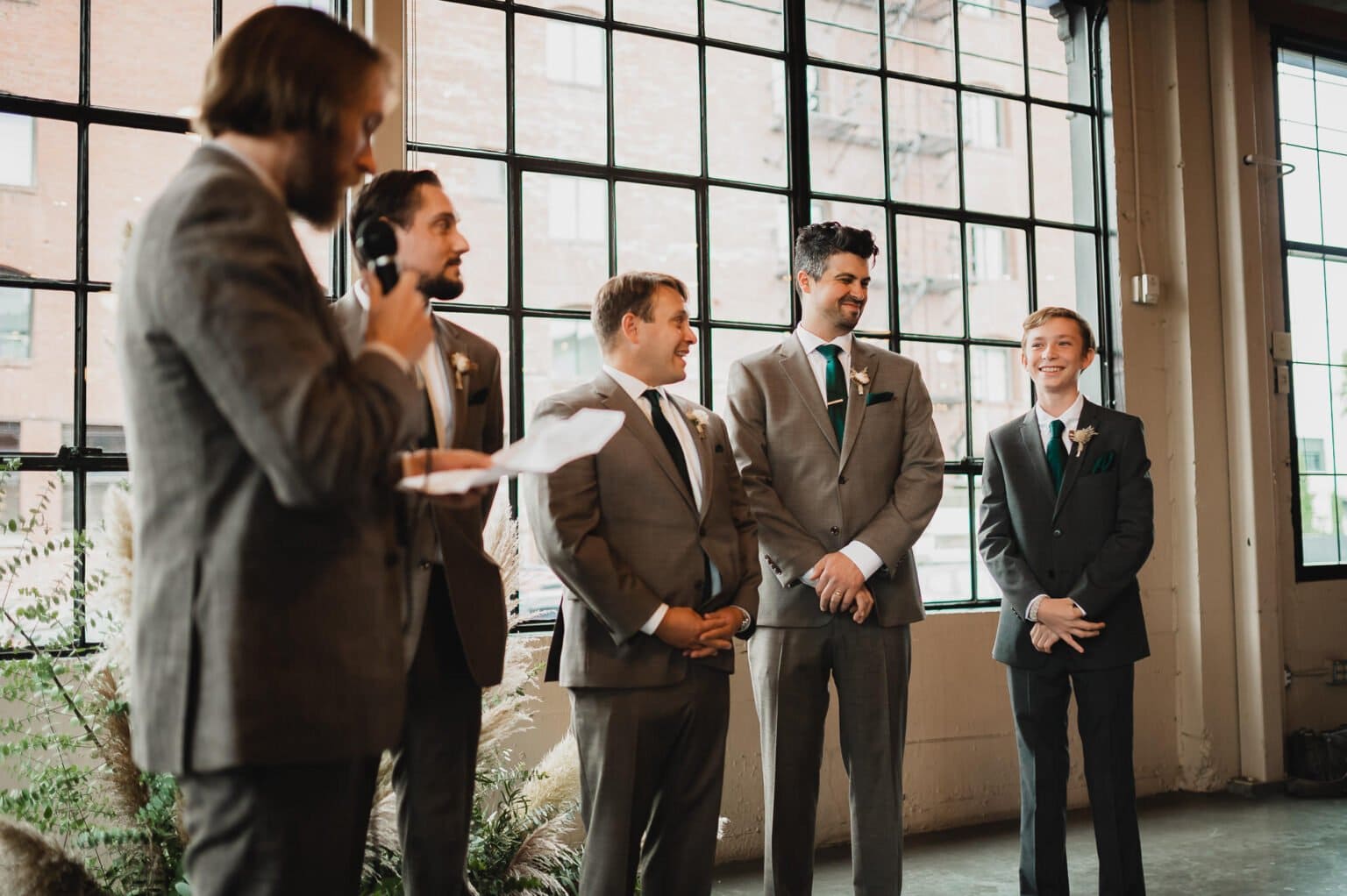 Four men in suits stand smiling indoors near large windows at Castaway Portland, as another man speaks into a microphone.