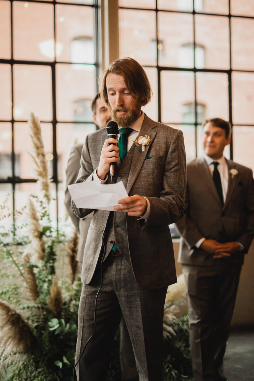 A man in a suit speaks into a microphone, holding paper, with others gathered near large windows. The setting offers a refined ambiance similar to capturing wedding photos at Castaway Portland.