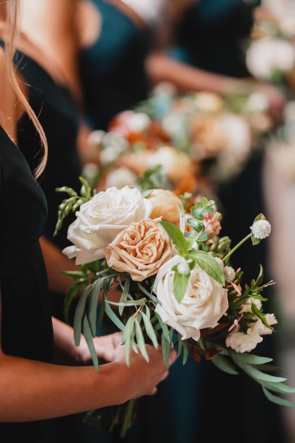 People lined up holding elegant bouquets filled with white and peach roses alongside lush greenery, all wearing dark dresses at a wedding photoshoot at Castaway Portland.