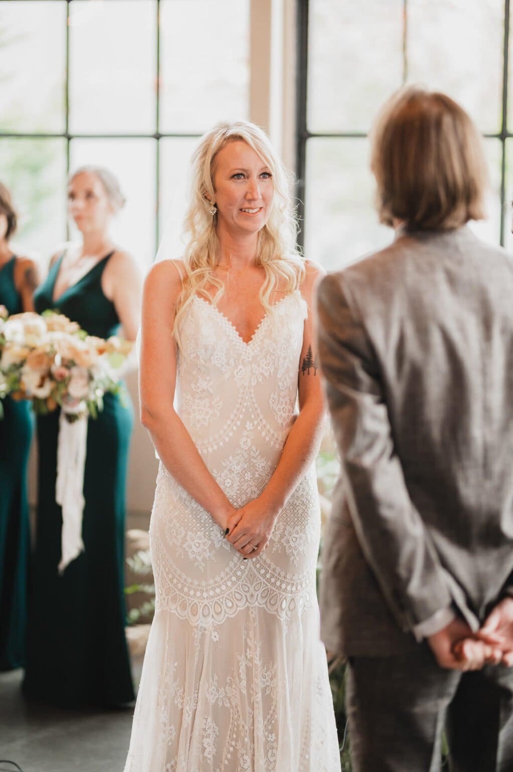 During the wedding ceremony, a bride in a lace gown faces her groom in a suit, with bridesmaids holding flowers behind. The elegant scene offers perfect wedding photos at Castaway Portland.