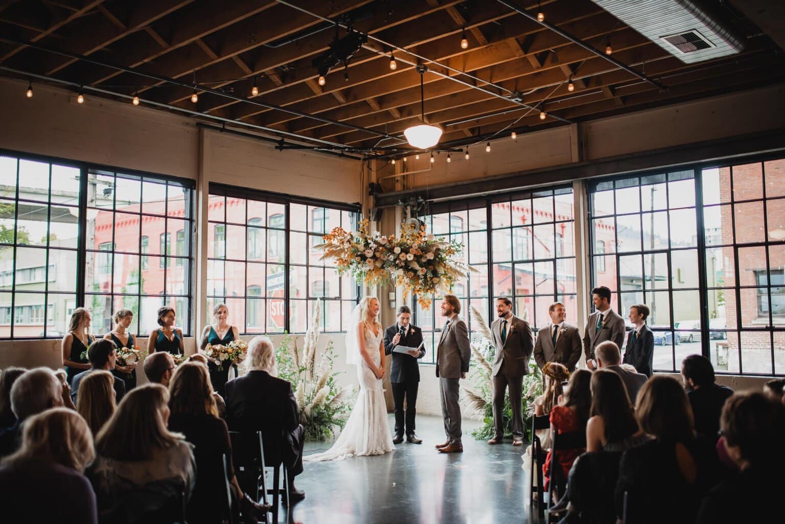 In an industrial-style venue with large windows and floral decorations, a couple exchanges vows while capturing stunning wedding photos at Castaway Portland.