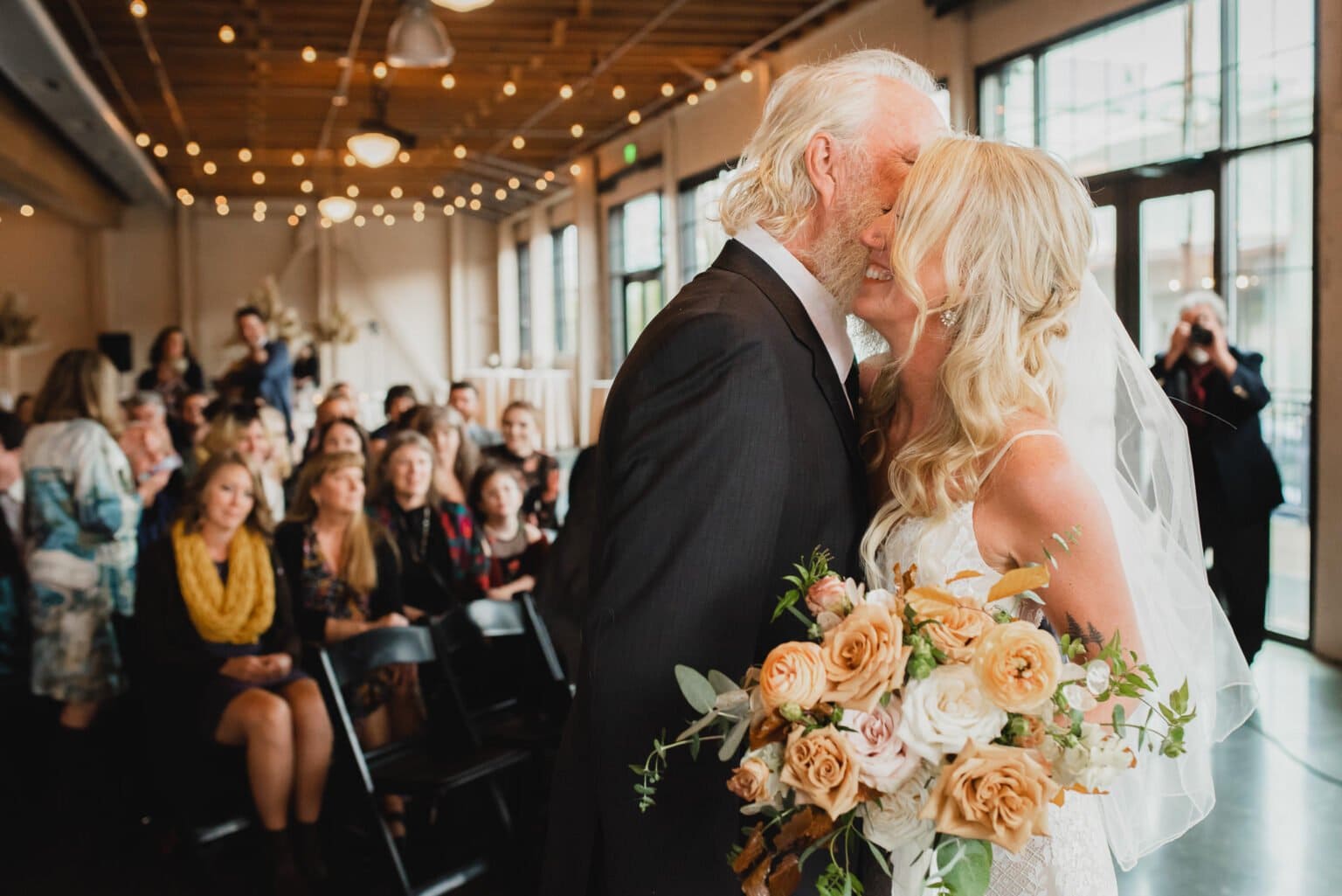 A man and woman share a kiss in a room filled with people, capturing a memorable moment reminiscent of wedding photos at Castaway Portland.