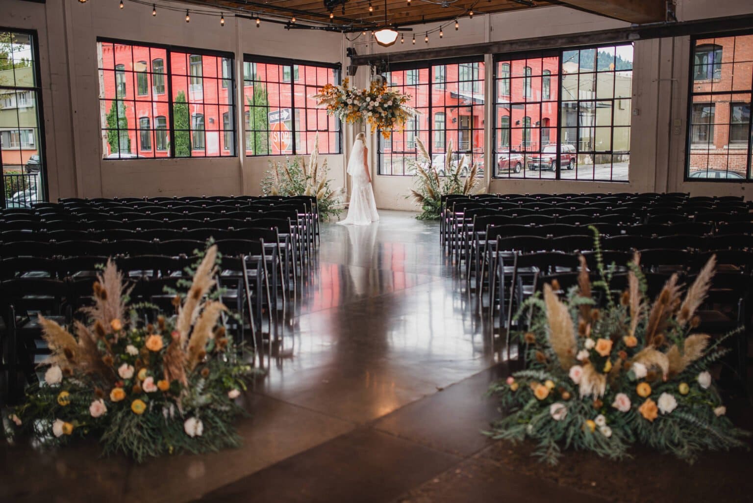 The bride stands near a floral arch in an empty, industrial-style wedding venue with black chairs and large windows, capturing stunning wedding photos at Castaway Portland.