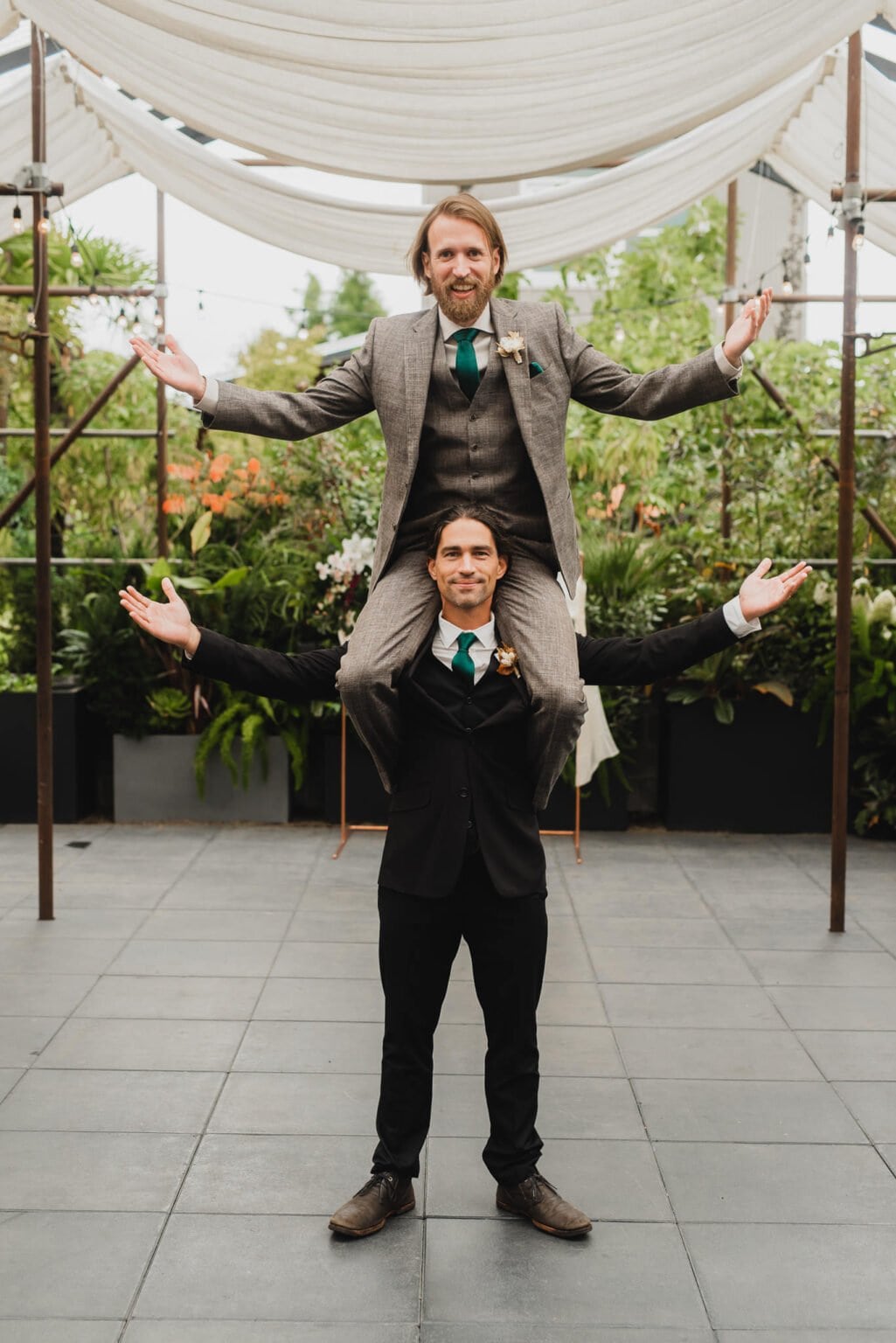 In a playful portrait at Blockhouse, two men in suits are captured striking a fun pose before a wedding at Castaway Portland; one man stands with his arms out while the other joyfully sits on his shoulders, mirroring the stance.