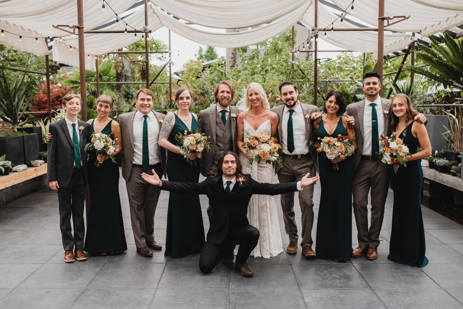 The bridal party gathers for portraits at Blockhouse before a wedding at Castaway Portland, surrounded by lush plants in the outdoor venue. One person kneels in front, arms spread wide.