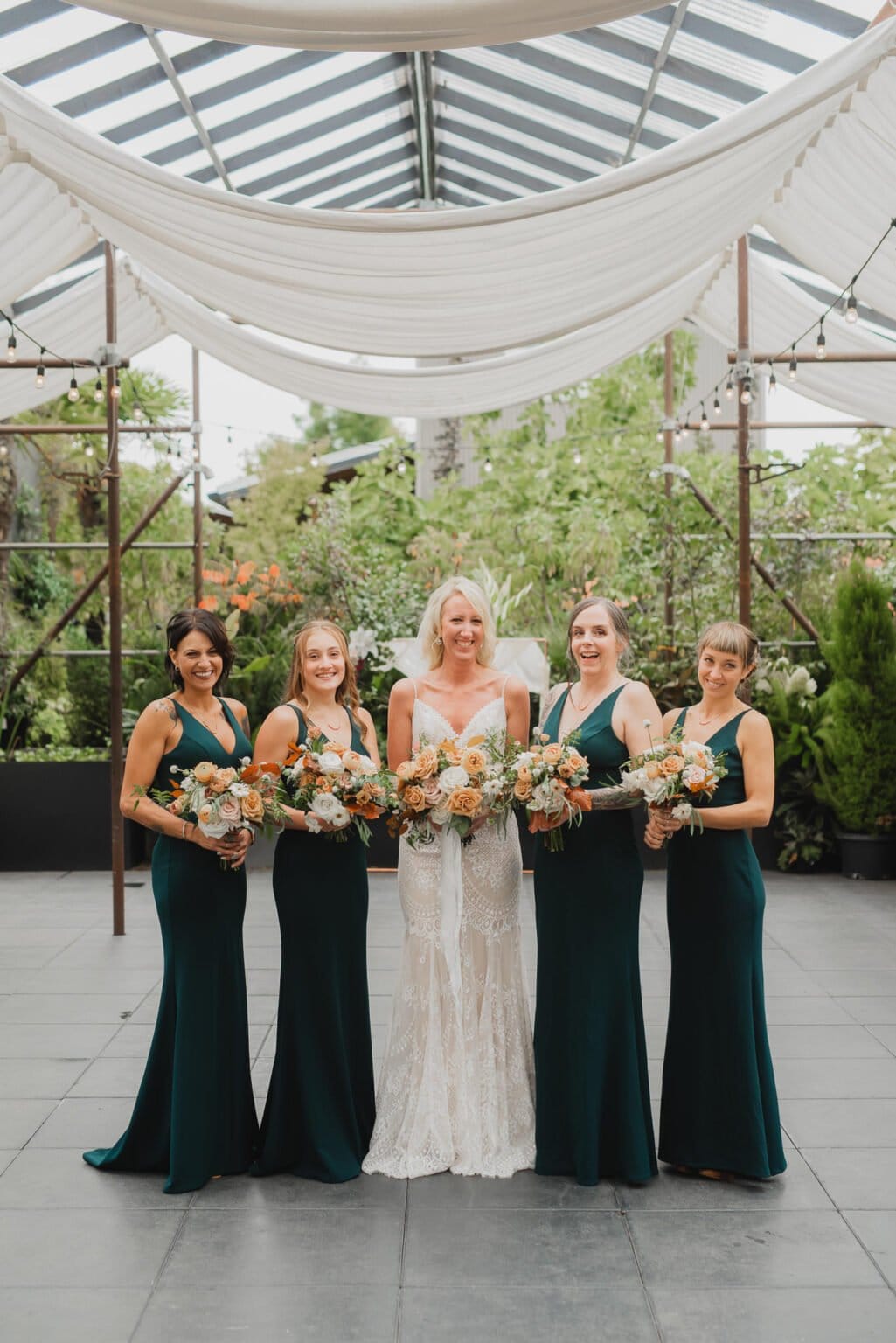 The bride in a white gown, accompanied by her four bridesmaids in green dresses, holds bouquets under a canopy. Their elegant portraits were captured at Blockhouse before a beautiful wedding at Castaway Portland.