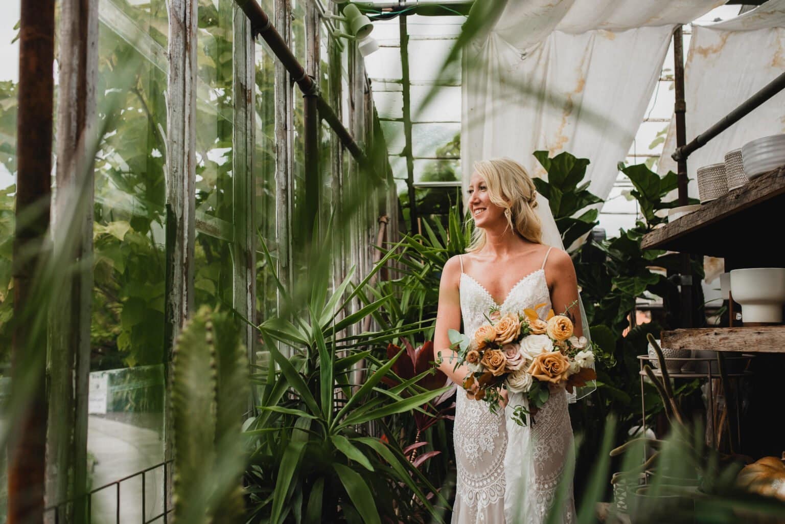 The bride, in a lace dress, stands gracefully in a lush greenhouse at Pomerius Nursery. She holds her bouquet under the soft light, capturing serene portraits before her wedding at Castaway Portland.
