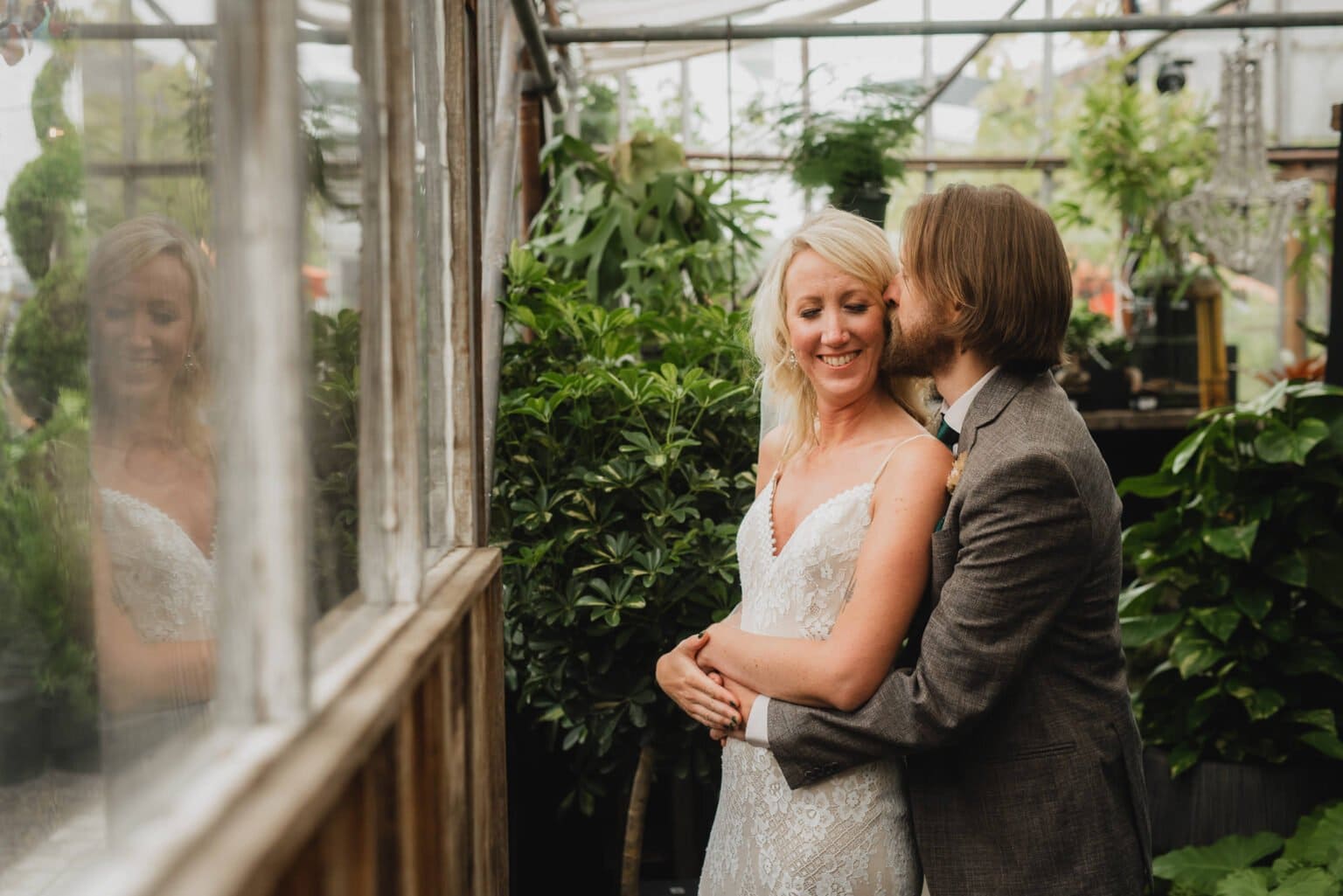 A couple embraces happily in a lush greenhouse at Pomerius Nursery, surrounded by vibrant green plants, as they prepare for their wedding at Castaway Portland.