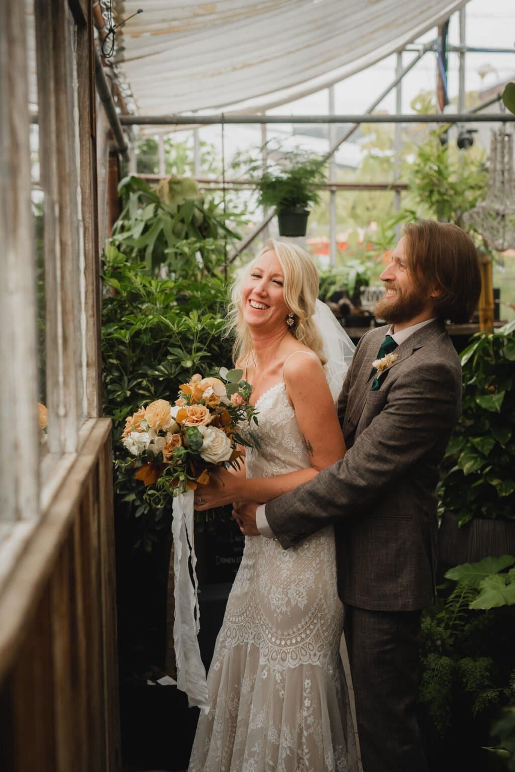 The bride in a lace dress and groom in a suit smile joyfully, standing together with a bouquet amidst the lush greenery of Pomerius Nursery, capturing stunning portraits before their wedding at Castaway Portland.