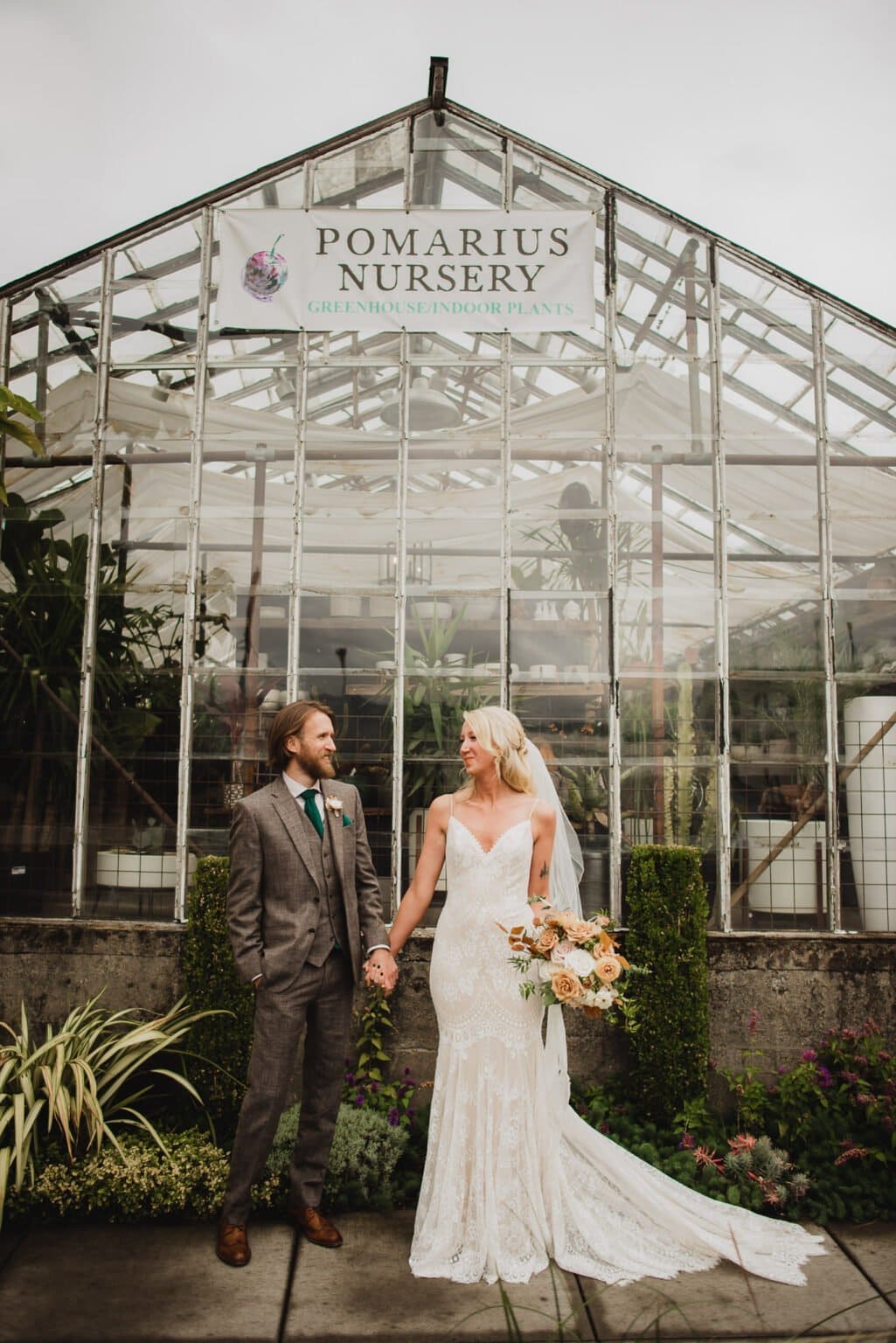 The bride and groom share a tender moment holding hands in front of a glass greenhouse labeled Pomarius Nursery, capturing beautiful portraits before their wedding at Castaway Portland.