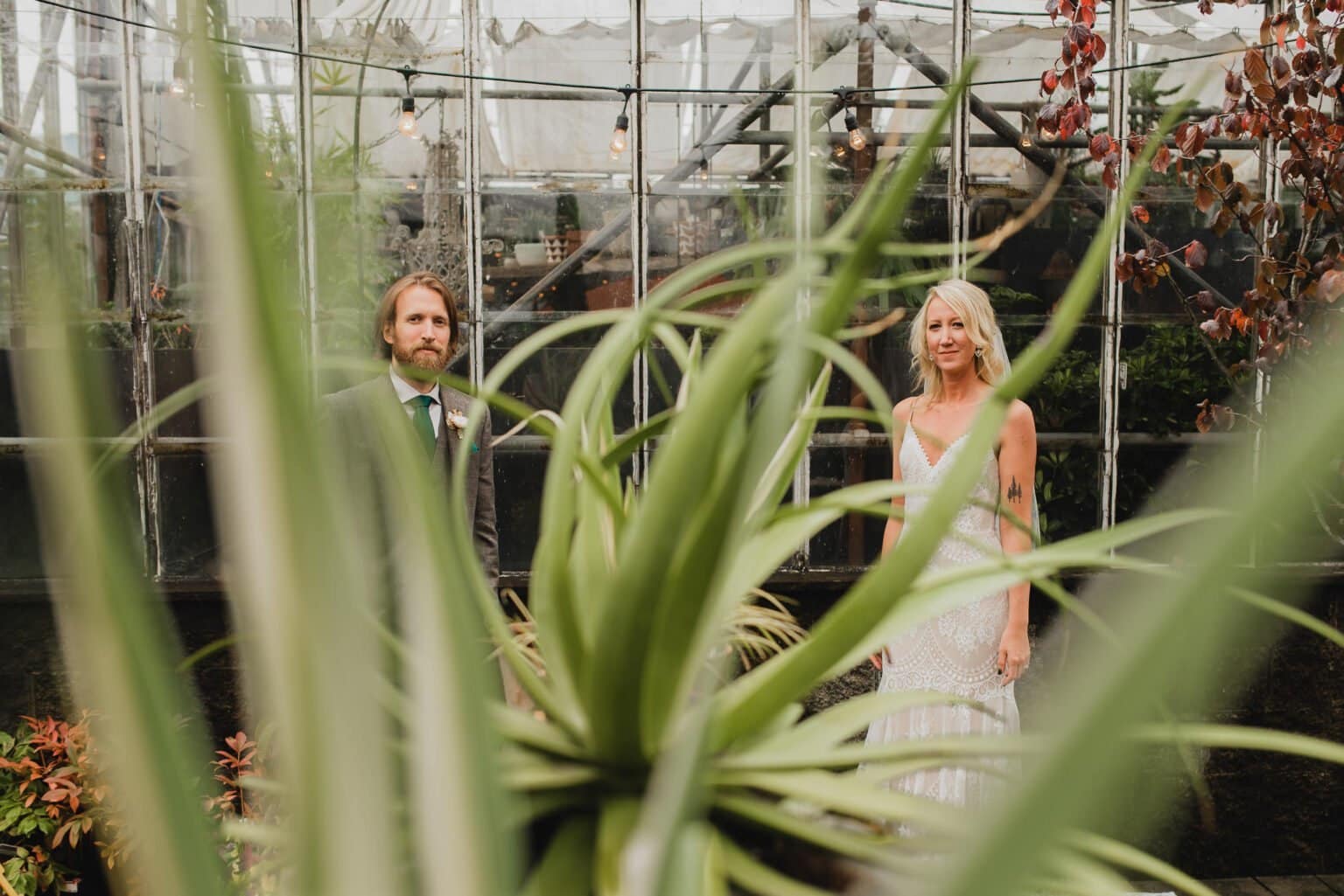 A couple poses in a greenhouse at Pomerius Nursery, partially obscured by the lush plant leaves in the foreground, just before their wedding at Castaway Portland.
