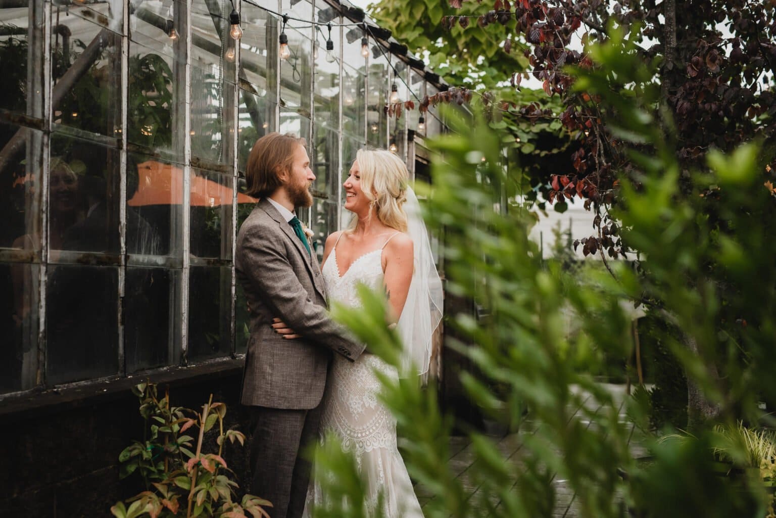 Bride and groom smiling at each other outside a glass greenhouse, surrounded by greenery, capturing beautiful portraits at Pomerius Nursery before their wedding at Castaway Portland.