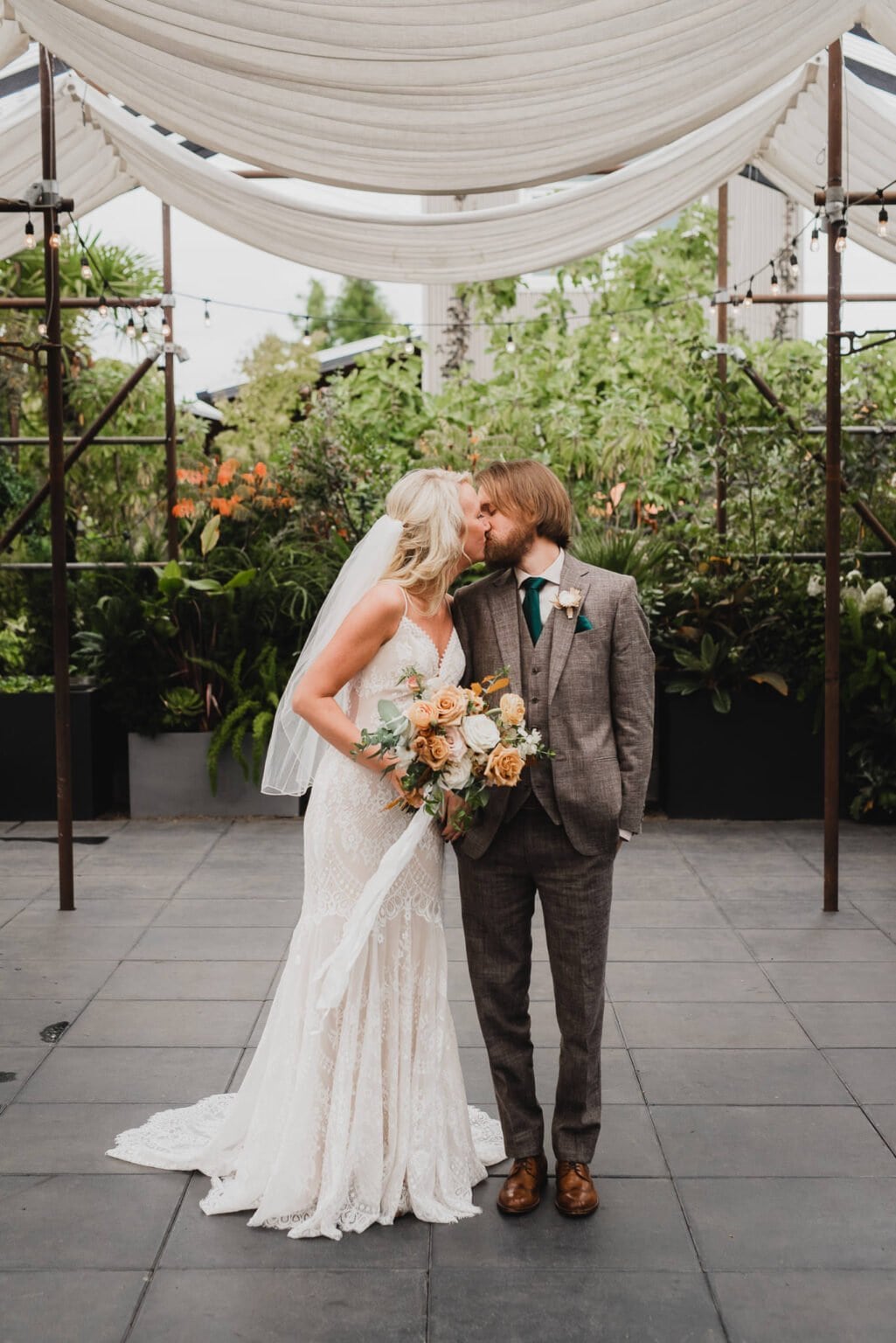 Bride and groom sharing a kiss under a canopy in a lush garden setting, both dressed in elegant wedding attire. Captured beautifully as part of their portraits at Blockhouse before their wedding at Castaway Portland.