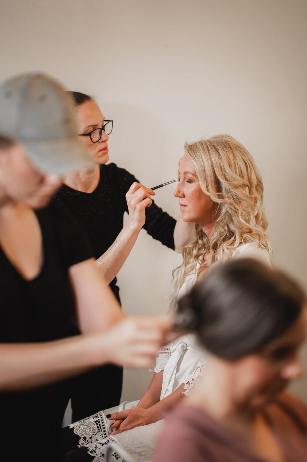 A makeup artist applies cosmetics to a seated woman with long blonde hair, getting ready at a nearby salon for a Castaway wedding, while another woman is visible in the foreground.