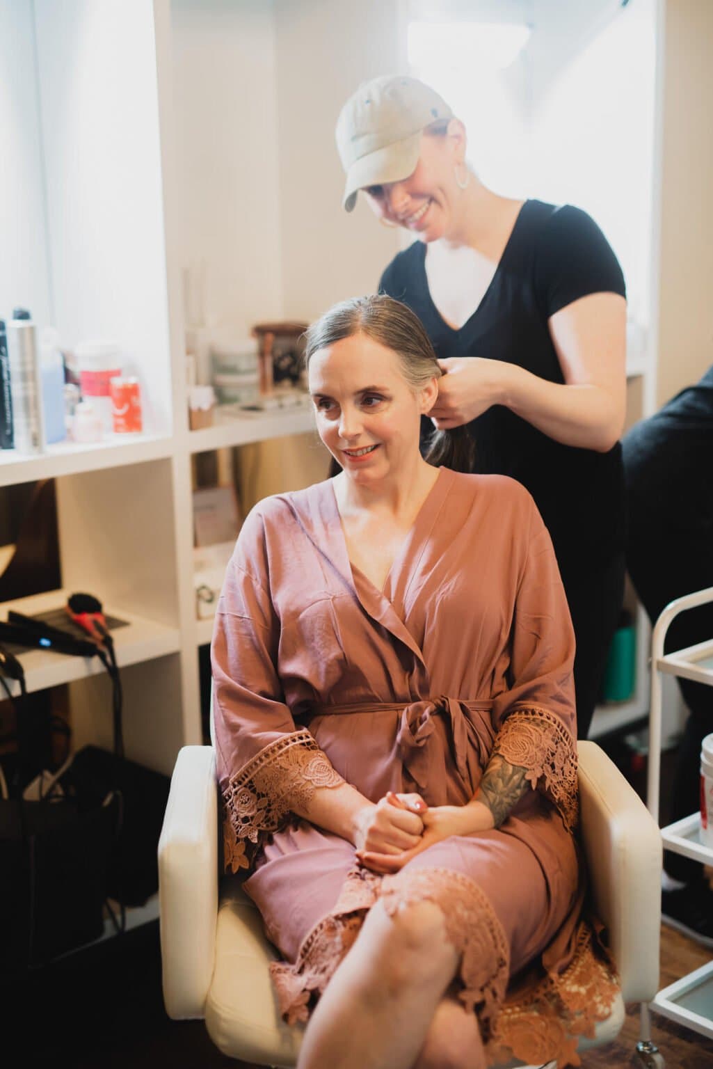 A bridesmaid in a pink robe is getting their hair styled by someone in a black shirt and cap, preparing at a nearby salon for a Castaway wedding.