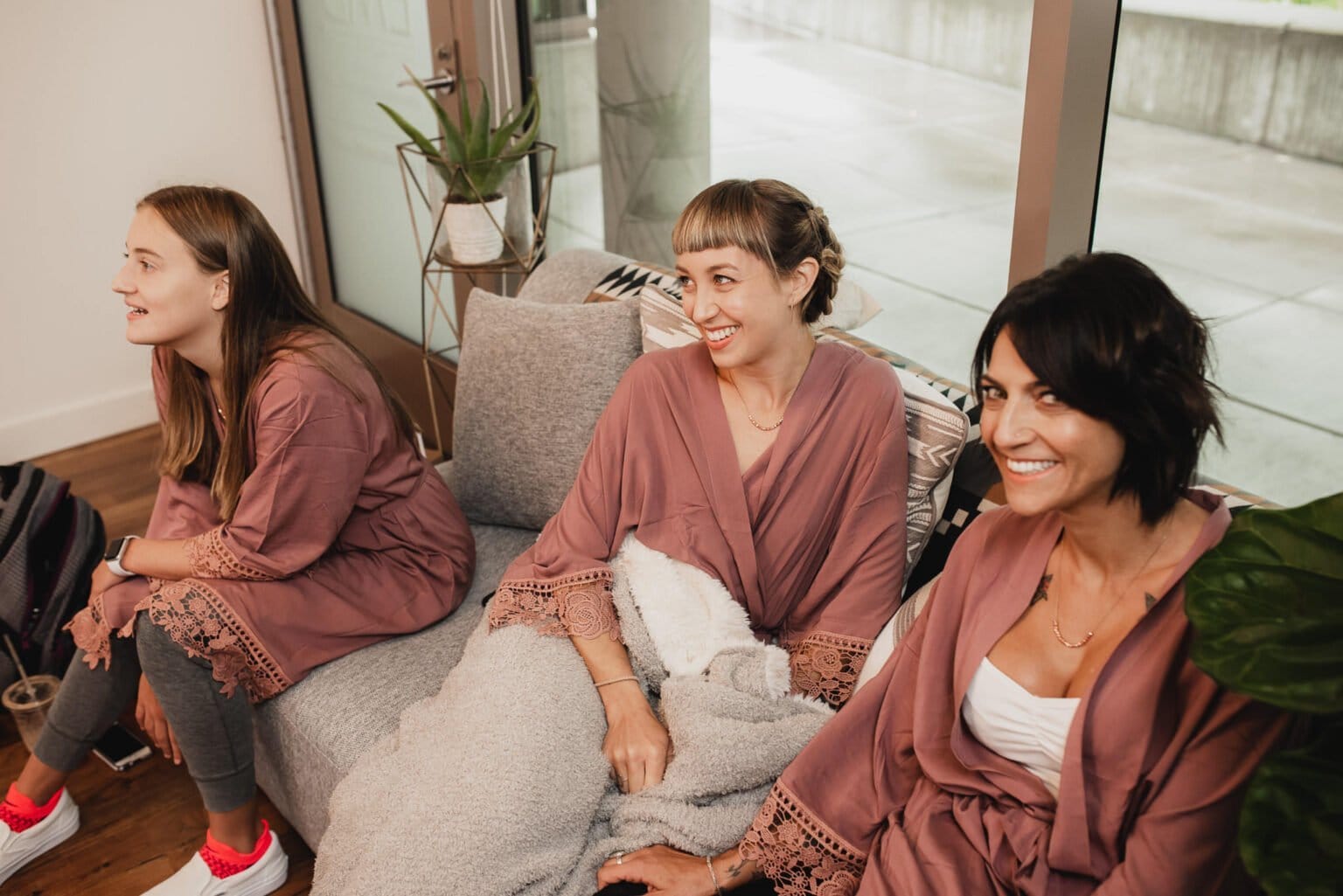 Three bridesmaids in matching robes sit and smile on a couch near a window with plants nearby, getting ready at a nearby salon during a Castaway wedding.