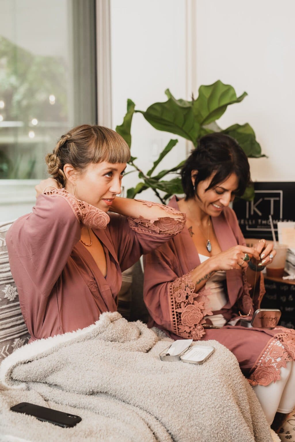 Two bridesmaids in pink robes are at a nearby salon, getting ready for a Castaway wedding. One adjusts her hair while the other smiles, looking down at a string of beads.
