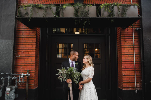 A wedding portrait of a couple just married holding hands and smiling at each other while standing in front of The Evergreen, a Portland indoor wedding venue.