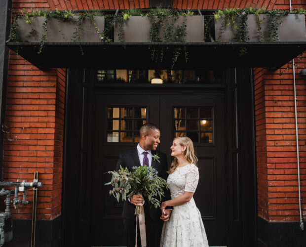A wedding portrait of a couple just married holding hands and smiling at each other while standing in front of The Evergreen, a Portland indoor wedding venue.