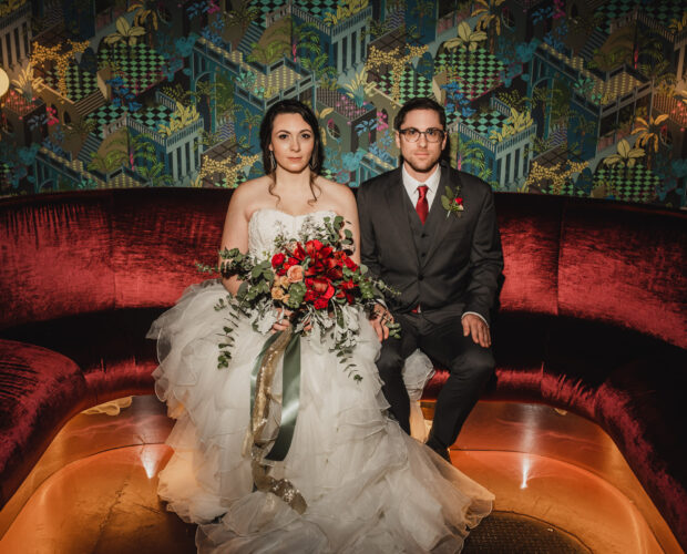 A wedding portrait of newlyweds sitting stoically on a sofa at Voysey, a speakeasy under their wedding venue, The Evergreen, in Portland, Oregon.