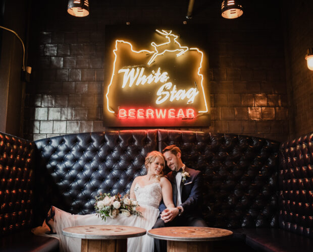 Newlyweds cuddle in a booth underneath a neon sign while having their wedding portraits taken at Loyal Legion, which is underneath the bride and groom's venue The Evergreen, one of Portland, Oregon's best indoor wedding venues.